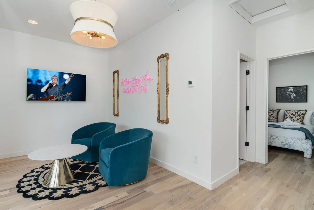 A modern living room with blue velvet chairs, a round coffee table, a TV displaying a performer, vibrant neon wall art, and framed artwork in an adjacent room. The room exudes a lively, social vibe ideal for group gatherings or entertainment.
