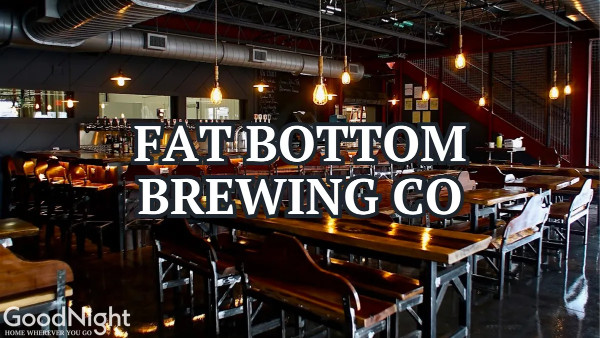 A spacious interior of a craft beer bar or brewery with long wooden tables, industrial lighting, and a visible bar area. Groups of people are gathered, socializing, tasting beers, and enjoying casual conversation in a friendly setting.