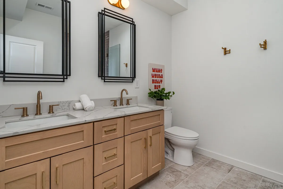 A modern bathroom with double vanity sinks, large mirrors, a decorative sign, and a potted plant. The space feels fresh and inviting, ideal for self-care routines and unwinding after a long day.