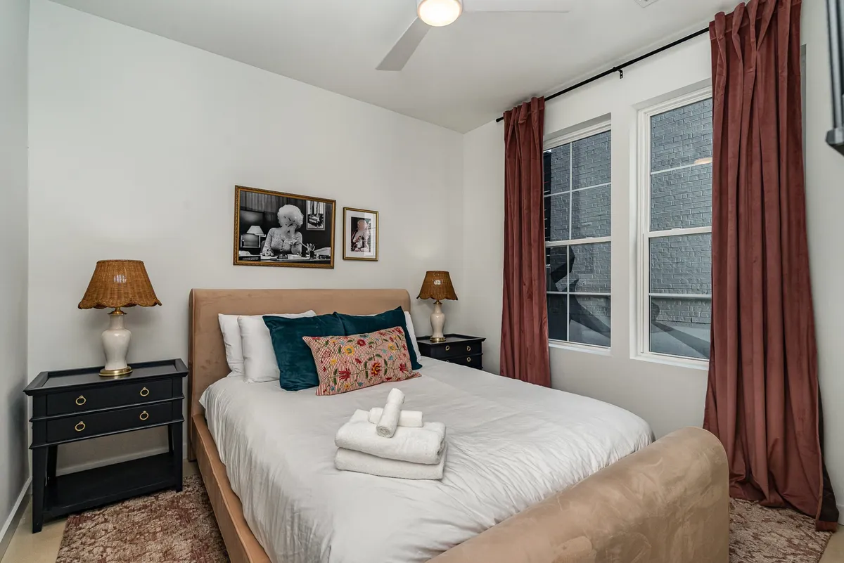 A stylish bedroom with a plush bed adorned with decorative pillows, complemented by artwork on the wall and nightstands with elegant lamps. The room exudes natural light, creating a cozy and welcoming atmosphere ideal for relaxation or reading.