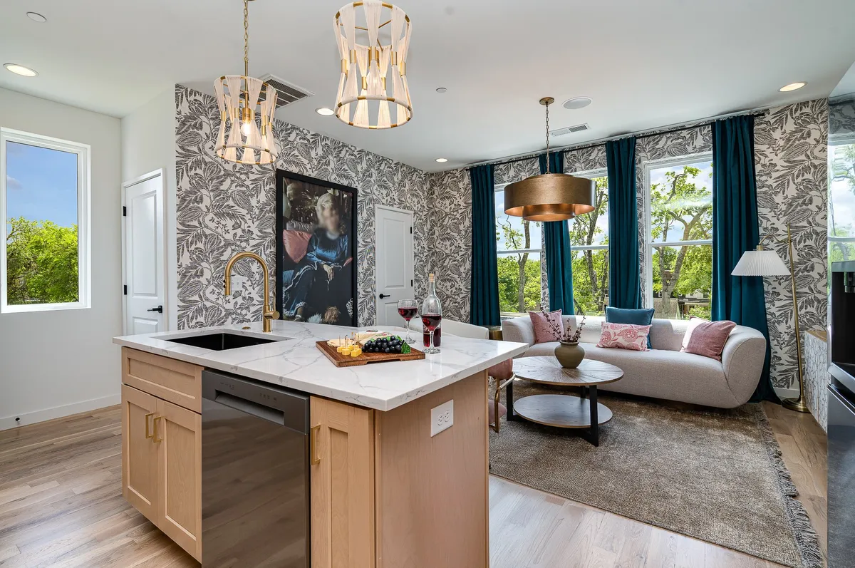 A stylish open-concept space that combines a modern kitchen with a spacious island, a comfortable sofa, contemporary artwork, and large windows offering views outside. The room glows with natural light and features inviting furnishings that encourage gathering and conversation.