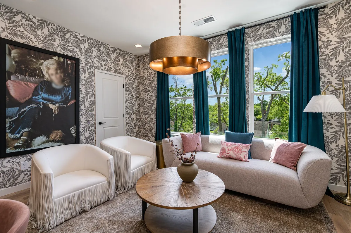 An elegant, comfortable lounge with stylish armchairs, a round wooden coffee table, wall art featuring a seated figure, and large windows offering a view of greenery. The ambiance is both social and tranquil, suitable for relaxation or intimate conversation.