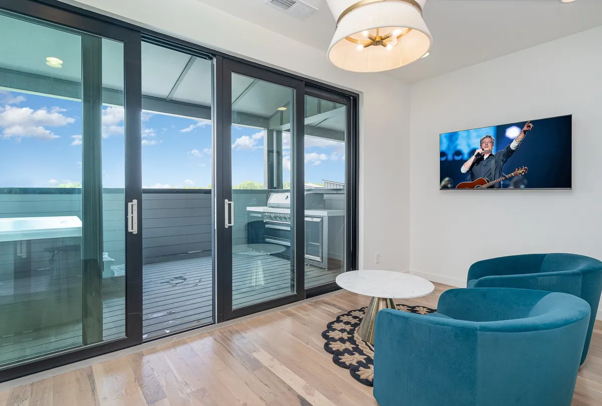 A contemporary living area featuring large glass sliding doors, blue armchairs, a round coffee table, and a television displaying a performance image. The space is filled with natural light and offers a view of the outdoors, promoting a sense of openness and flexibility.
