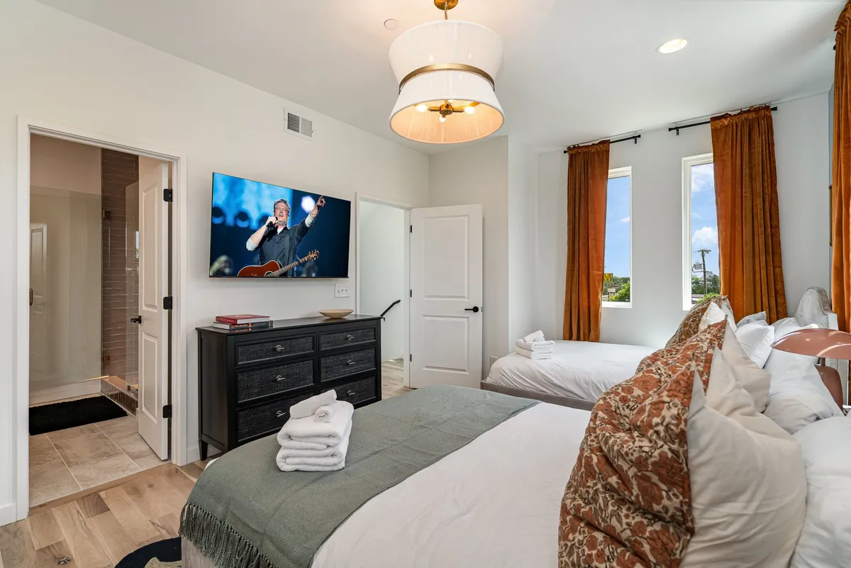 A bright and inviting bedroom featuring two beds with decorative pillows, a large TV, and windows with curtains. The space is filled with natural light and provides a cozy, flexible sleeping arrangement ideal for groups or families.