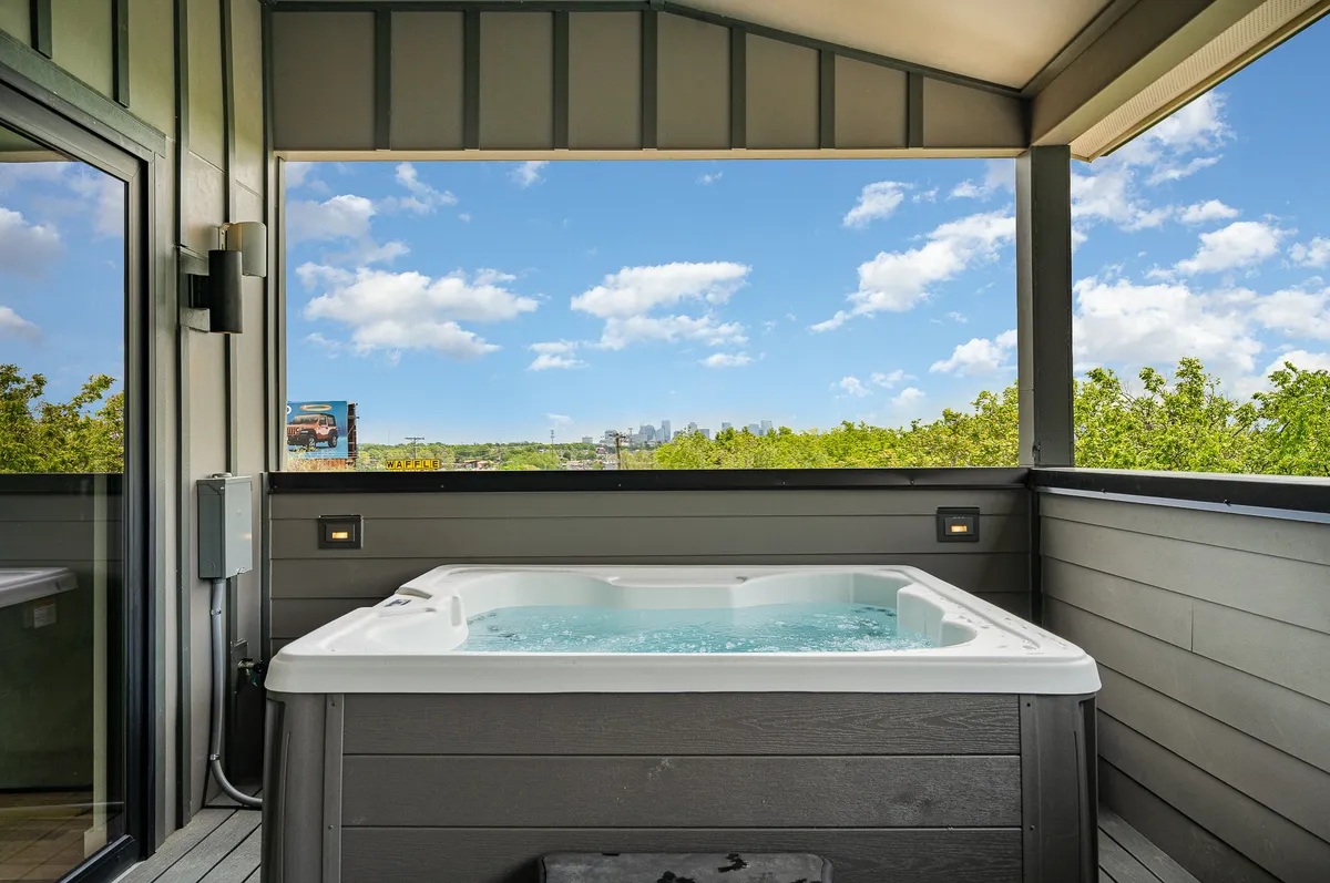 A private deck is featured with a luxurious hot tub, offering a sweeping view of the city skyline, framed by green trees and set under an open blue sky. The inviting atmosphere suggests a perfect spot for soaking, socializing, and enjoying memorable sunsets or evenings under the stars.