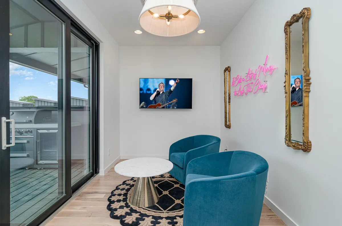 A chic living area with two blue chairs, a round coffee table, a TV displaying a musical performance, a neon sign, and mirrors. The atmosphere is vibrant yet cozy, ideal for relaxing or enjoying a movie or music night with friends or family.