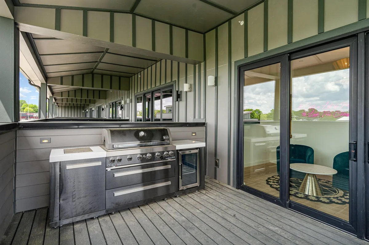 The deck is shown with a sleek outdoor grill, comfortable sitting area, and large glass doors leading inside. The setup is ideal for group barbecues, al fresco dining, and easy indoor-outdoor transitions, matching the festive and social atmosphere depicted in the section.