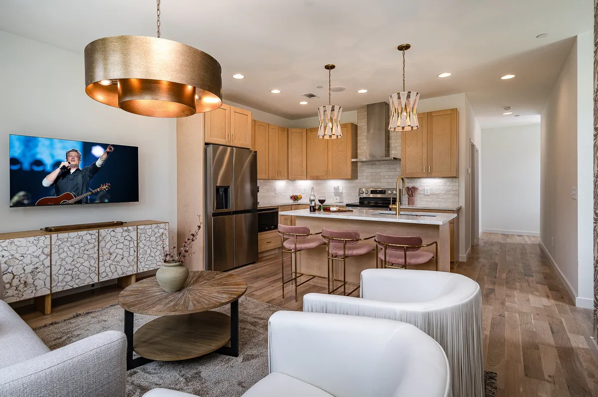 A stylish, open-concept living space featuring a modern kitchen island with bar stools, a cozy seating area centered around a unique coffee table, and a large television displaying a live performer, evoking the spirit of Nashville. The setup is perfect for entertaining, relaxing, or gathering with friends and family.